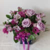 Lavender roses and pink flowers arranged in a square glass vase with a ribbon.