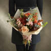Handheld bouquet of peach, red, and pink flowers wrapped in paper