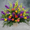 Large arrangement of purple and yellow flowers with orange roses in a white vase