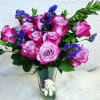 Pink roses and blue filler flowers in a clear glass vase with a small teddy bear