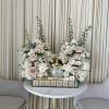 Two pastel floral arrangements flank a gold bottle on a table