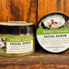 Two jars of herbal infused facial scrub on a wooden surface.