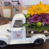 Flower arrangement in a white pickup truck planter with a sunflower and purple daisies
