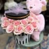 Pink roses arranged in a striped gift box with a teddy bear behind it