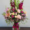 Tall mixed bouquet of pink, peach, and yellow flowers with green spikes in a clear vase with a magenta bow