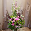 Tall pink and green flower arrangement with roses and delphinium in a clear glass vase on a wooden table.