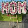 Large floral standing tribute spelling "MOM" in pink and white flowers