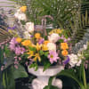 Arrangement of yellow and white flowers with a glass cross, set among tall palm greenery on a white pedestal.