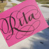 A pink sign with the name "Rita" held outdoors on a suburban street.