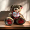 Brown teddy bear with red sequin heart reading "I Love You" on a wooden surface