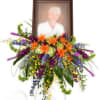 Standing funeral spray with orange lilies and purple flowers around a framed portrait