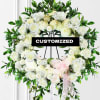 White floral wreath on a stand with green foliage and a pink ribbon