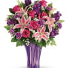 Pink lilies, roses, and purple flowers arranged in a tall purple glass vase
