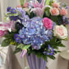 Pastel bouquet with hydrangea, roses, lilies, and purple filler flowers in a lavender vase