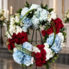 Heart-shaped wreath of red roses, blue and white hydrangeas, and white flowers on a stand