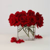 Red roses arranged in a clear square glass vase with scattered petals