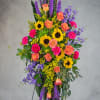Tall standing spray of sunflowers, orange roses, pink carnations, and purple blooms on an easel.