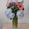 Mixed bouquet with blue hydrangeas, pink gerbera daisy, and purple blooms in a clear vase