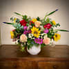 Colorful mixed arrangement with sunflowers, roses, and daisies in a white ceramic vase on a wooden table.
