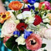 Colorful hand-tied bouquet with orange ranunculus, blush roses, red blooms, and blue accent flowers.