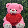 Pink teddy bear holding a red heart against a green hedge backdrop