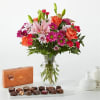 Mixed bouquet of pink lilies, roses, and daisies in a glass vase with chocolates