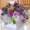 Purple shades mixed with green flowers in white box. Adorable centerpiece arrangement