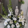 This sympathy arrangement contains a ceramic figure of Madonna adorned with fresh