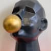This unique sculpture features a matte black geometric face blowing a gold