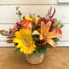 Orange roses, sunflowers, and sunset lilies feature in this arrangement with rust