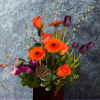 This striking arrangement features a vibrant blend of orange gerbera daisies, purple
