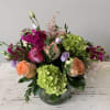Low mixed arrangement of peach and pink roses, green hydrangeas, and purple blooms in a clear round vase