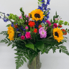 Vibrant mixed bouquet with sunflowers, pink and orange blooms, and blue delphinium in a clear glass vase.