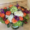 Low round arrangement of orange roses, green and white hydrangeas, and purple blooms in a glass vase