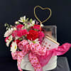 Pink and white flower arrangement with a heart topper and gift items in a round box