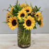 Sunflowers beautifully arranged in a glass vase with seasonal greenery. Stands approximately