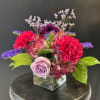 Low arrangement of pink and purple flowers with a lavender rose in a square glass vase.