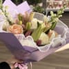 Hand-tied bouquet of white and peach roses with pink lilies, wrapped in lavender paper with ruffled white trim