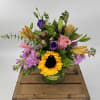 Mixed arrangement of sunflowers, pink roses, lilies, and purple blooms in a round glass vase