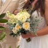 Bridal hand-tied bouquet of pale yellow roses and cream flowers with soft greenery, held by a bride in white.