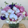 Round arrangement of white orchids, hydrangeas, and pink and purple roses in a glass vase.