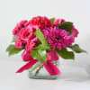 Bright pink roses, carnations, and a gerbera daisy in a square glass vase with a pink ribbon bow.