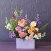 Colorful mixed flower arrangement in a lavender gift bag-style container against a purple wall