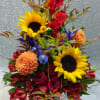 Tall mixed arrangement with sunflowers, red roses, orange dahlias, and purple iris in a clear glass vase