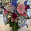 Mixed birthday arrangement with pink gerbera, blue delphinium, hydrangeas, and calla lilies in a clear vase.