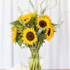 Tall bouquet of bright yellow sunflowers in a clear glass vase on a wooden surface.