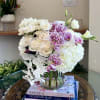 White roses, hydrangeas, and cascading orchids with purple speckled blooms in a clear glass vase on stacked books.