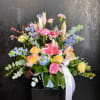 Pastel arrangement of pink lilies, roses, carnations, and blue flowers in a white vase with a white ribbon.