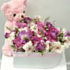 Pink teddy bear hugging a white hat box filled with pink and white flowers