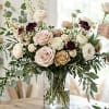 Large blush and cream rose arrangement with burgundy accents in a clear glass vase on a table